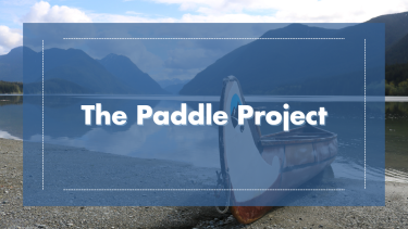 Scenic image of water and mountains with a canoe at shore. Blue overlay with title "The Paddle Project".